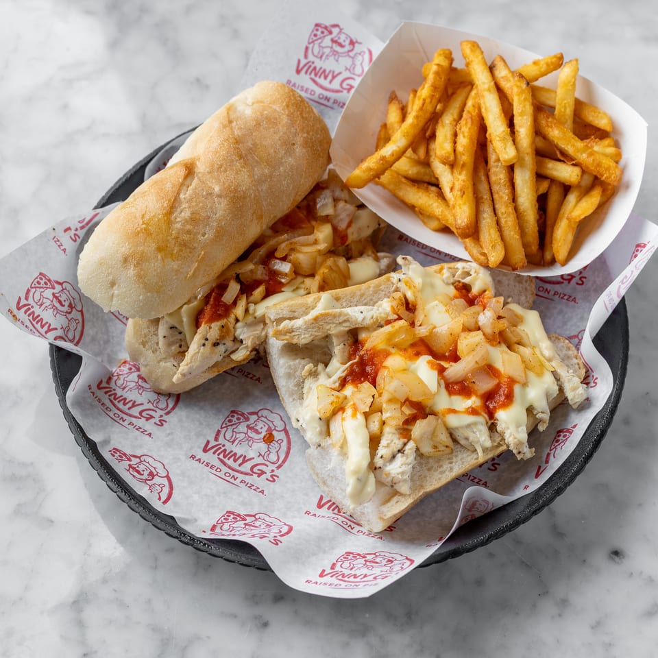Chicken cheesesteak with onions and sauce, served with fries on Vinny G's branded paper.