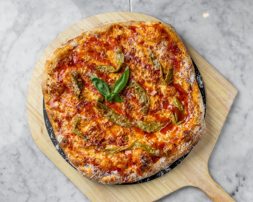 A whole pizza with red sauce, cheese, green peppers, and basil on a wooden peel.
