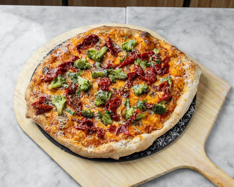 A freshly baked pizza with broccoli, sun-dried tomatoes, and cheese on a wooden peel.