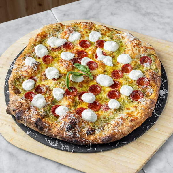 A pesto pizza with pepperoni, fresh mozzarella, and basil on a wooden pizza peel.