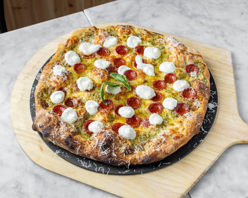 A pesto pizza with pepperoni, fresh mozzarella, and basil on a wooden pizza peel.