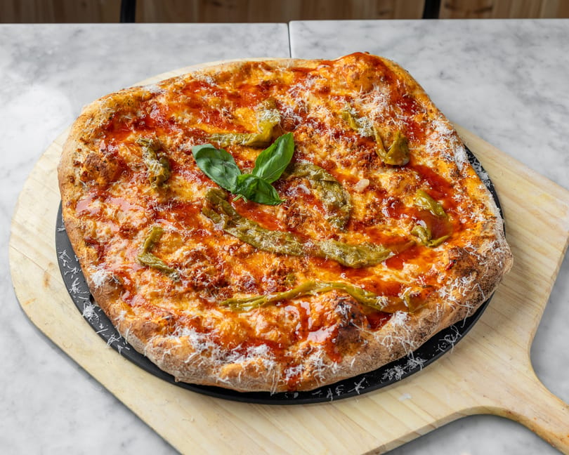 A freshly baked pizza with red sauce, melted cheese, green peppers, and basil on a wooden peel.
