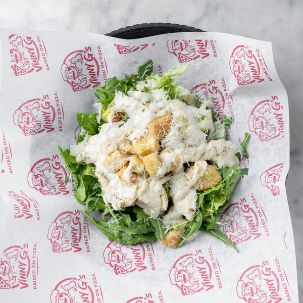 A Caesar salad with chicken, croutons, and dressing, served on Vinny G's branded paper.