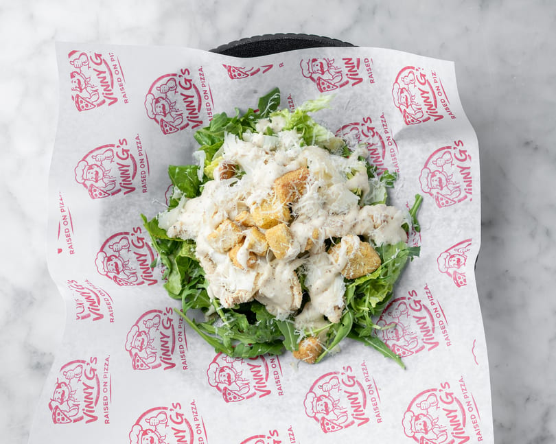 A Caesar salad with chicken, croutons, and dressing, served on Vinny G's branded paper.