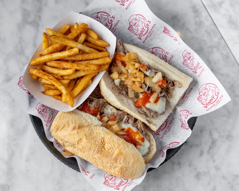Two Vinny G's cheesesteak sandwiches with onions and sauce, served with a side of fries.