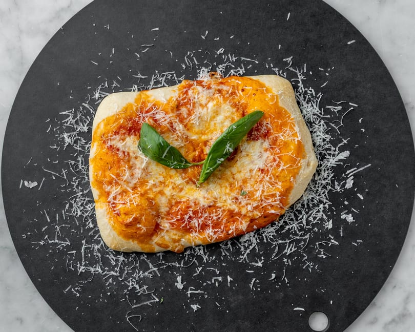 Rectangular flatbread pizza with tomato sauce, melted cheese, grated parmesan, and basil leaves on a black plate.