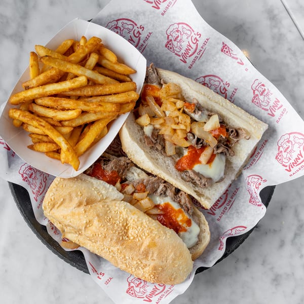 Two Vinny G's cheesesteak sandwiches with onions and red sauce, served with a side of fries.