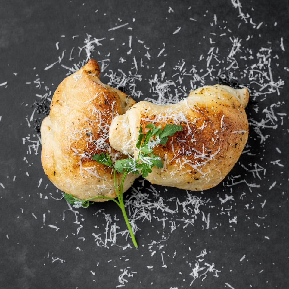 Two golden-brown calzones, sprinkled with Parmesan cheese and fresh parsley, on a dark gray surface.