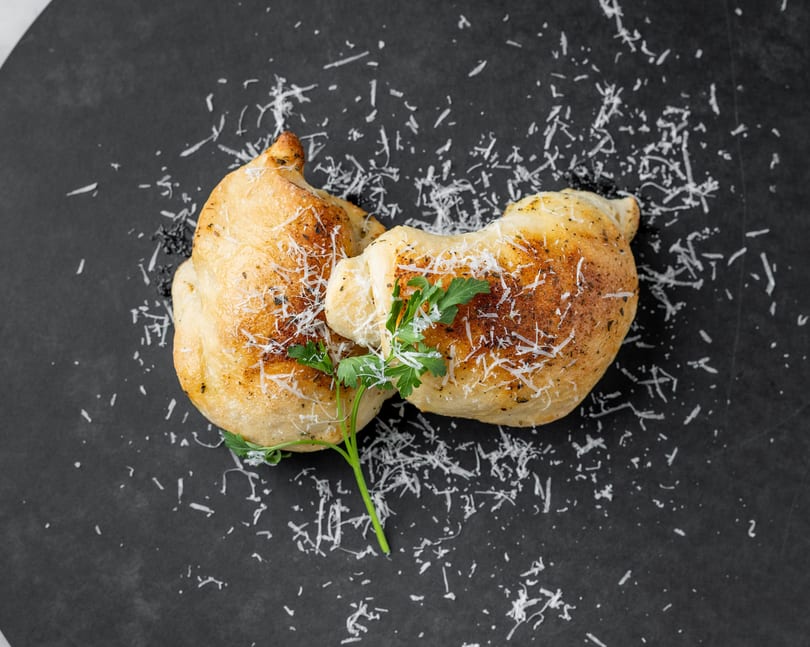 Two golden-brown calzones, sprinkled with Parmesan cheese and fresh parsley, on a dark gray surface.