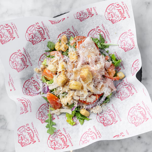 A fresh salad with chicken, tomatoes, red onions, croutons, and parmesan cheese on Vinny G's branded paper.