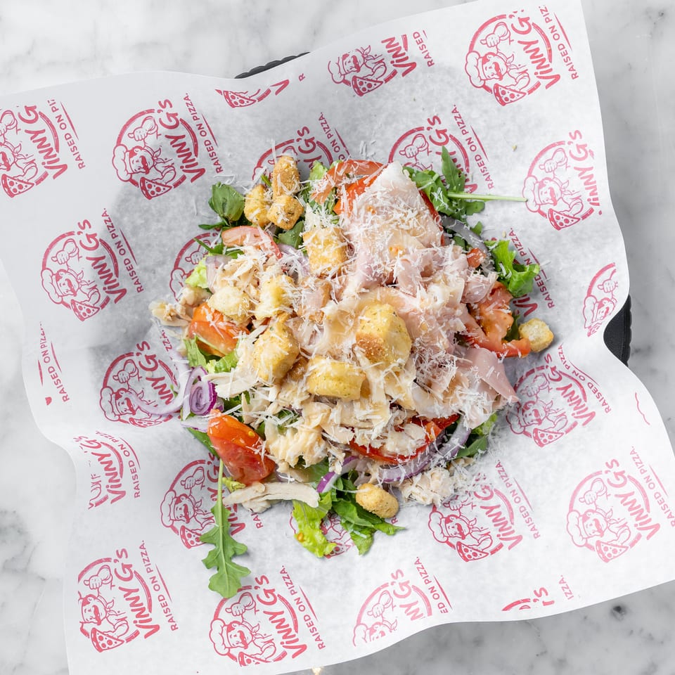 A fresh salad with chicken, tomatoes, red onions, croutons, and parmesan cheese on Vinny G's branded paper.