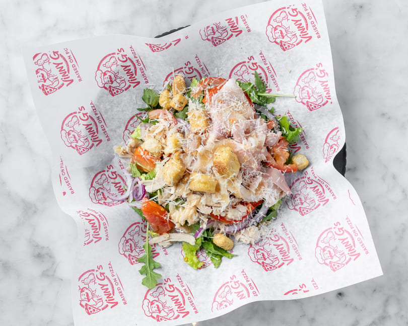 A fresh salad with chicken, tomatoes, red onions, croutons, and parmesan cheese on Vinny G's branded paper.